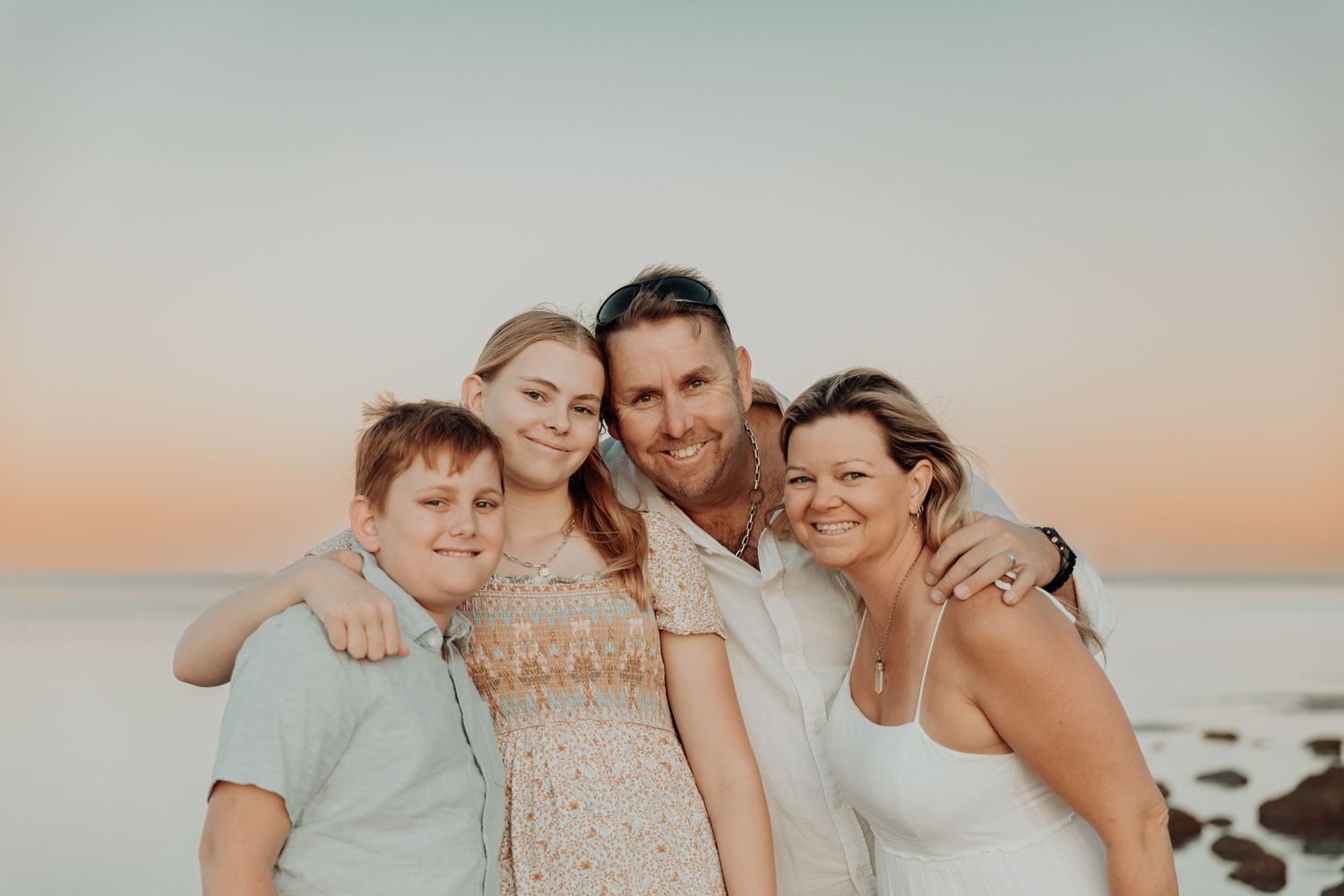 A family of four poses together on the beach at sunset with soft warm lighting and ocean waves in the background. Located in hearsons Cove,