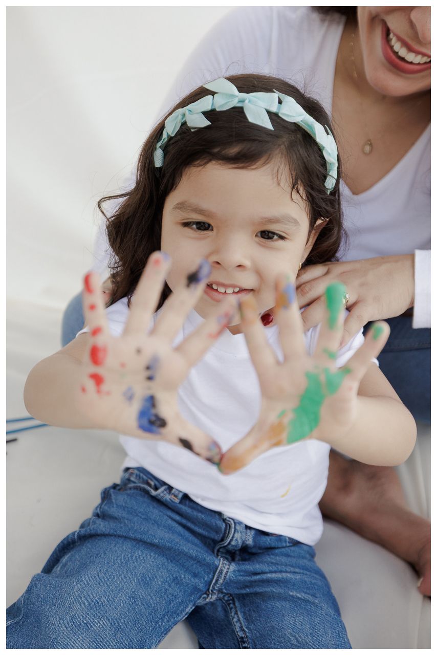 A small child shows their paint-covered hands while wearing denim and a white shirt with a mint hair bow.