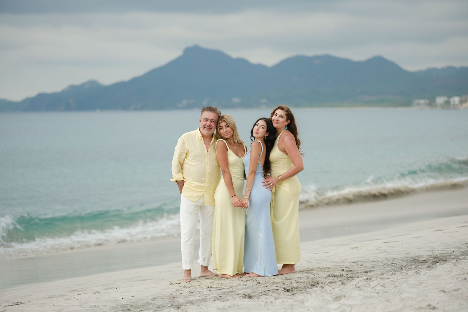 People in yellow and white clothing walk along a scenic tropical beach with mountains in the background.