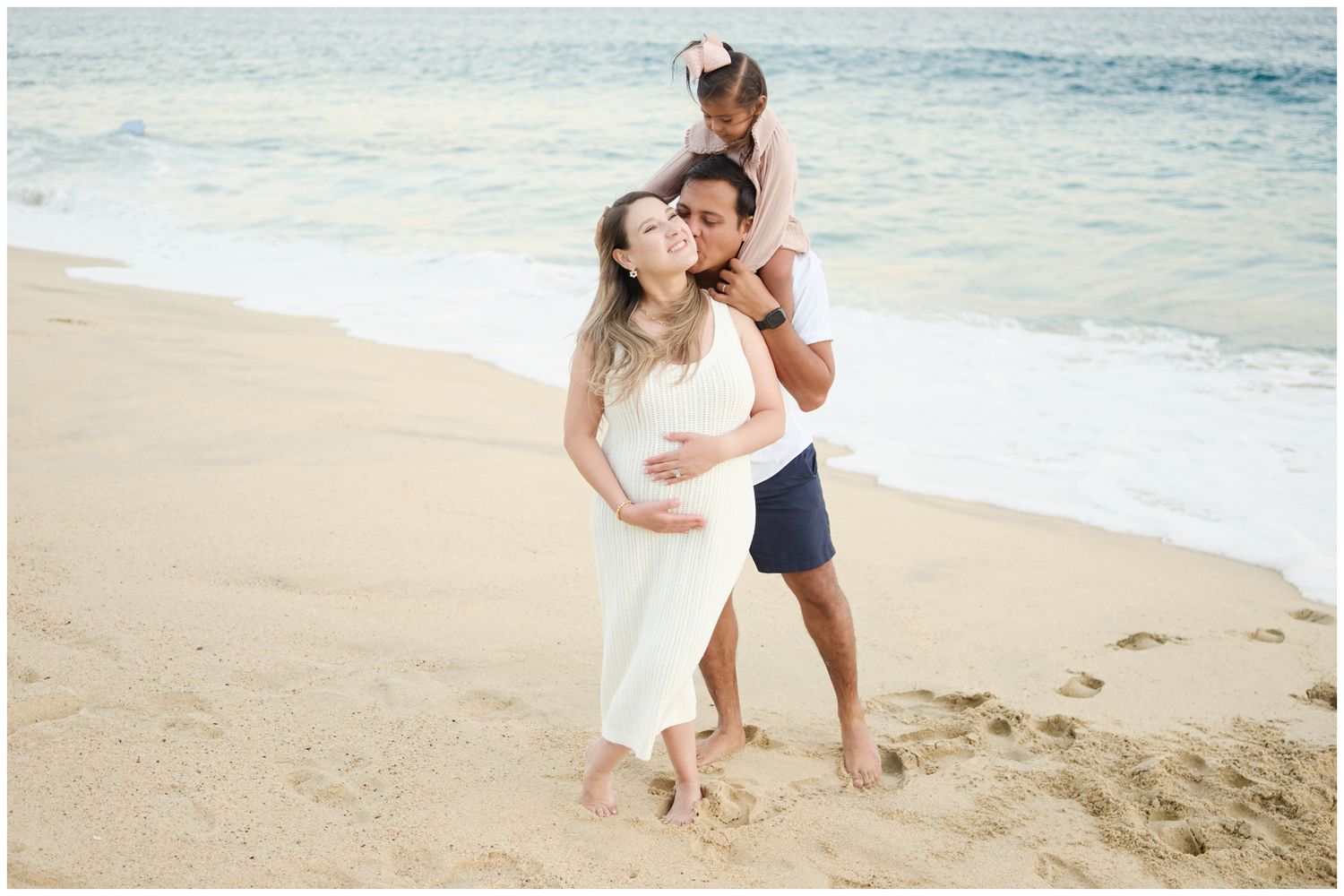 A couple enjoys a tender moment together on a sandy beach with gentle waves rolling in behind them.