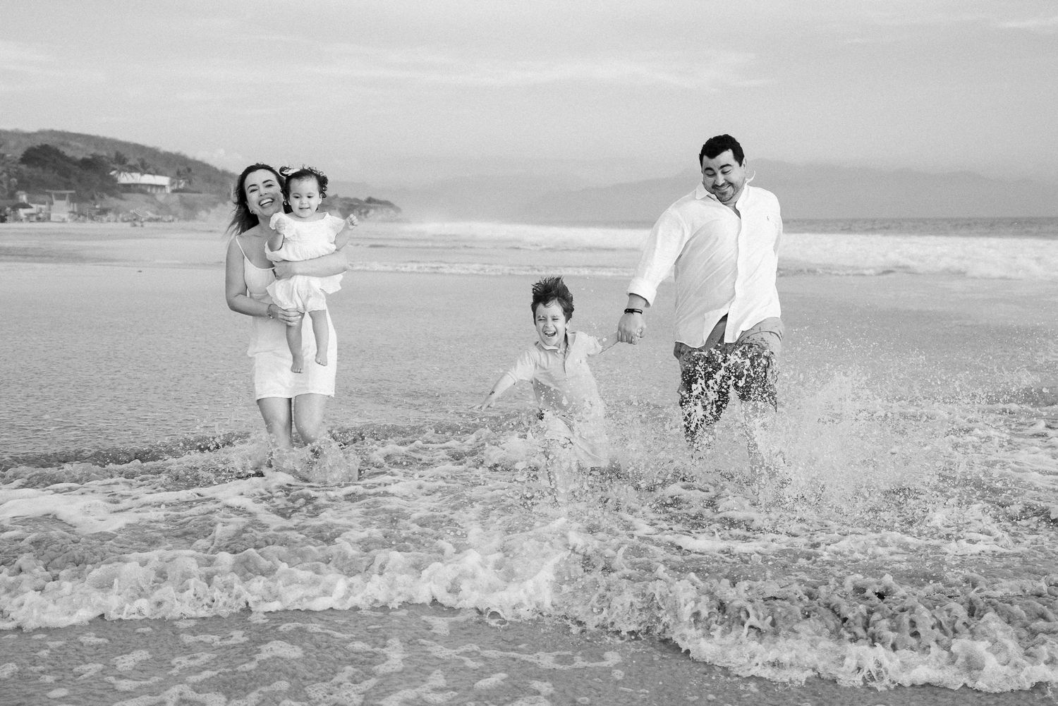 Family enjoying waves splashing at beach shore in black and white photograph.