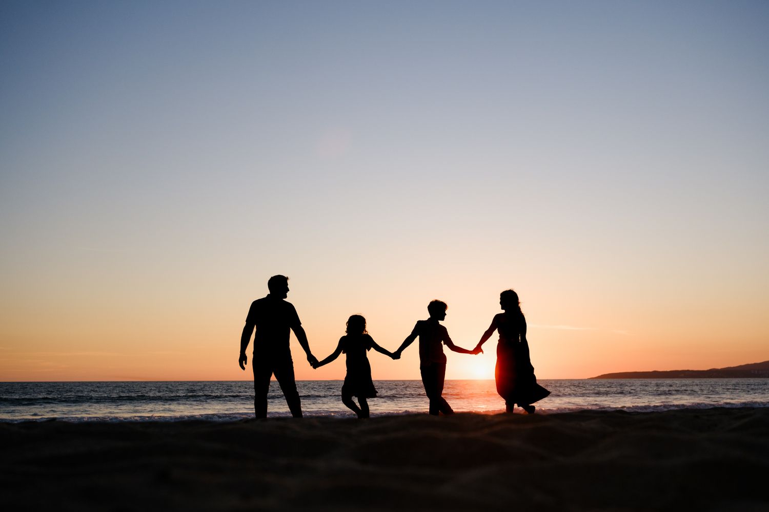 Silhouetted figures hold hands along a beach at sunset with an orange sky reflecting on the ocean.
