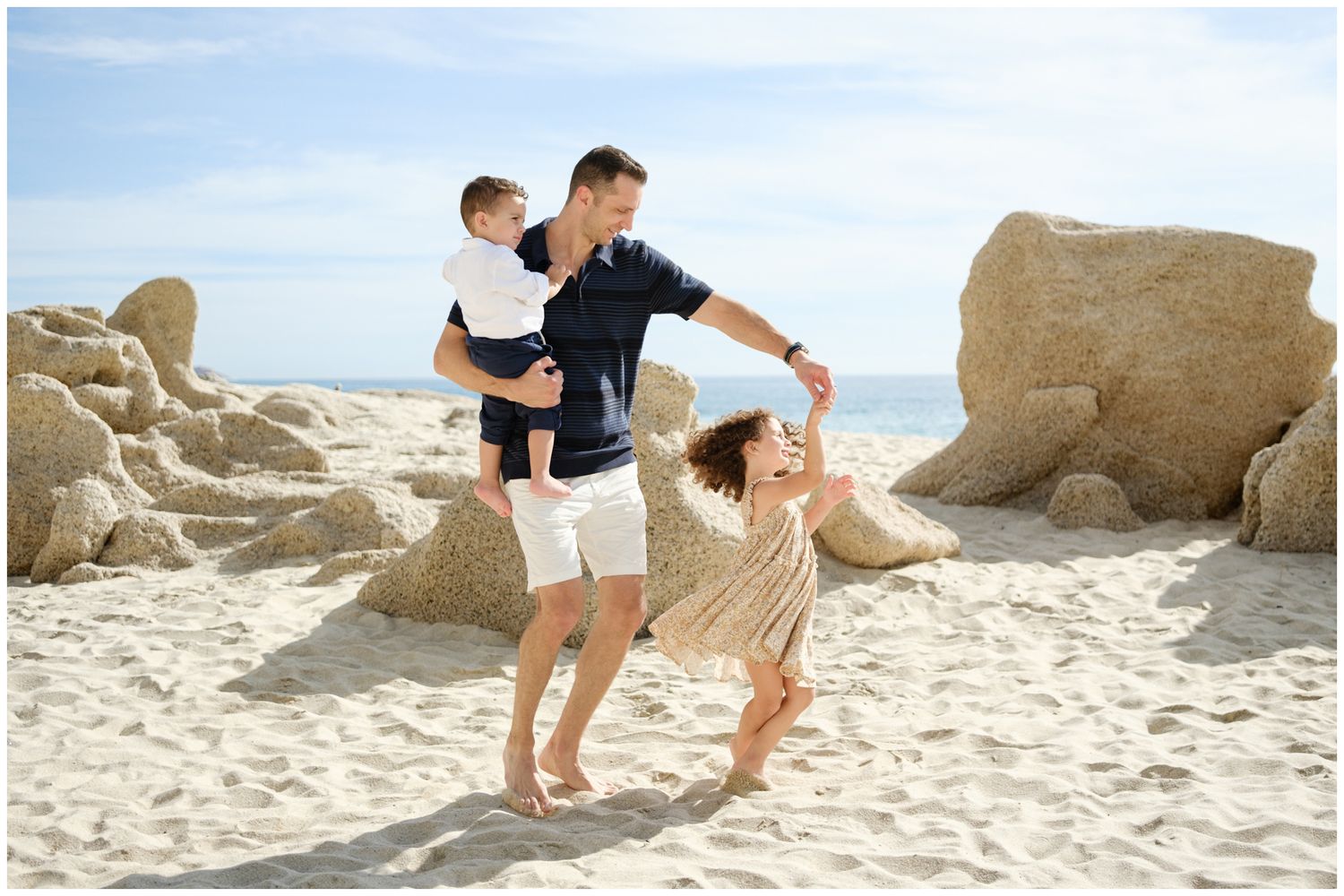 Parent and child play together on a sunny beach with rock formations in the background.