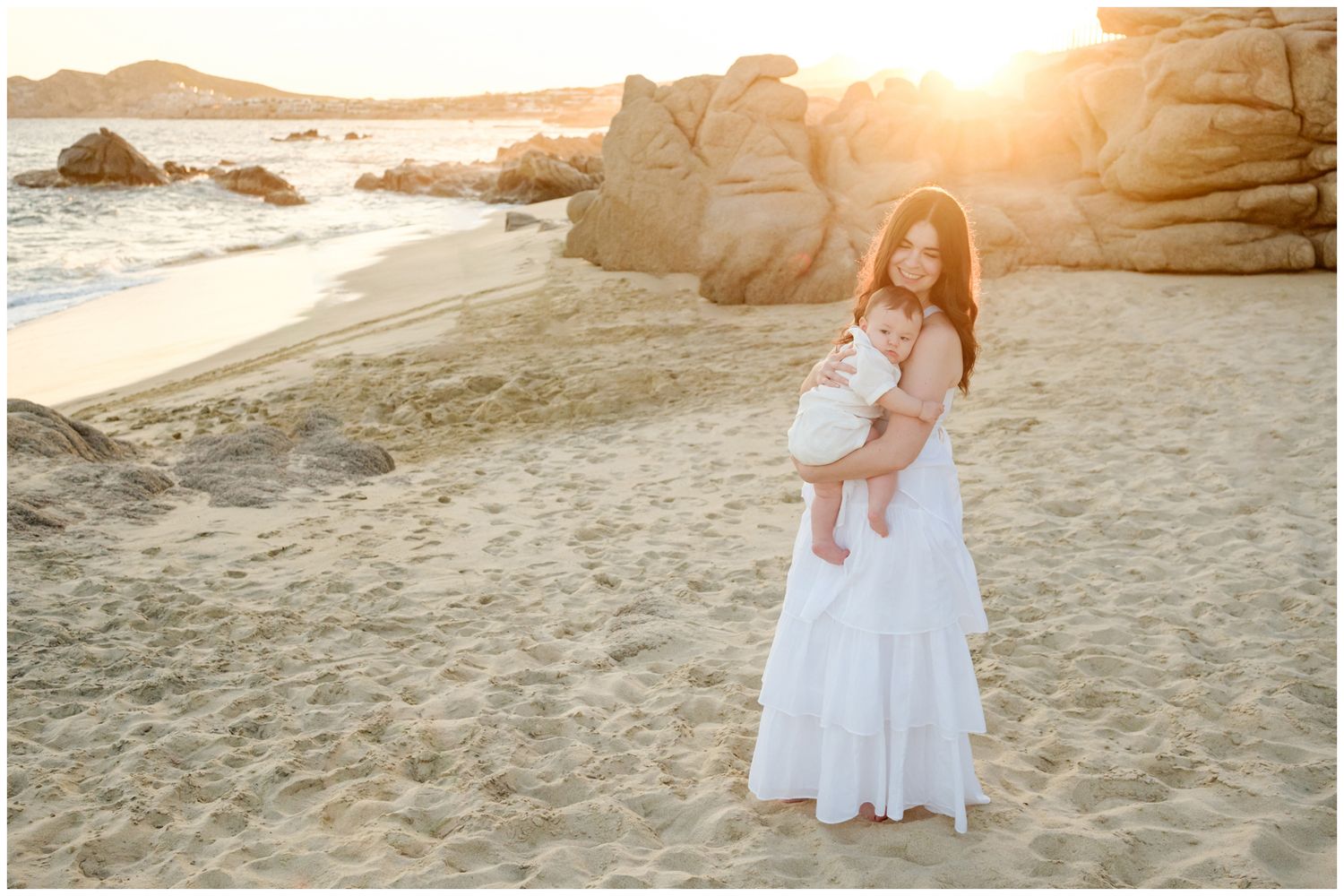 A figure in a flowing white dress holds a baby on a beach at sunset with rocky coastal formations in the background.