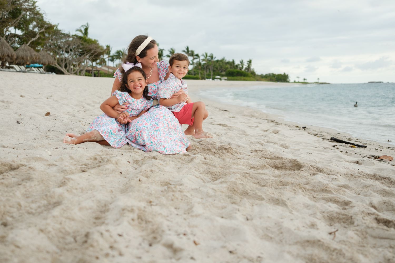 A family sits close together on a tropical beach with palm trees and ocean views in the background.