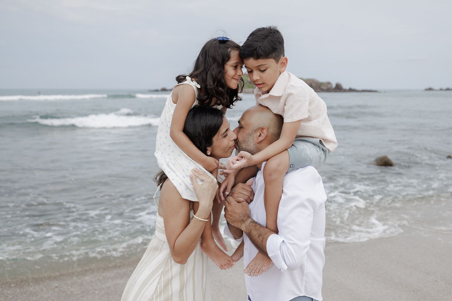 Family of four sharing a tender moment on a beach, parents holding their two children with ocean waves in the background.