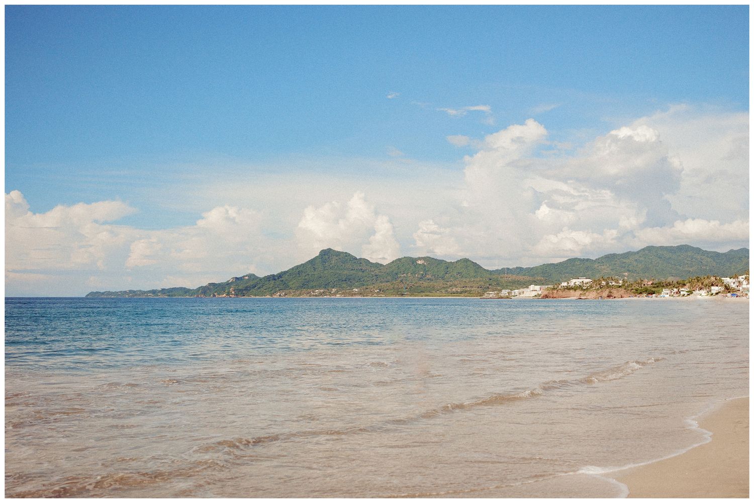 Beautiful ocean waves lap against a sandy beach with mountains and coastal buildings visible in the distance.