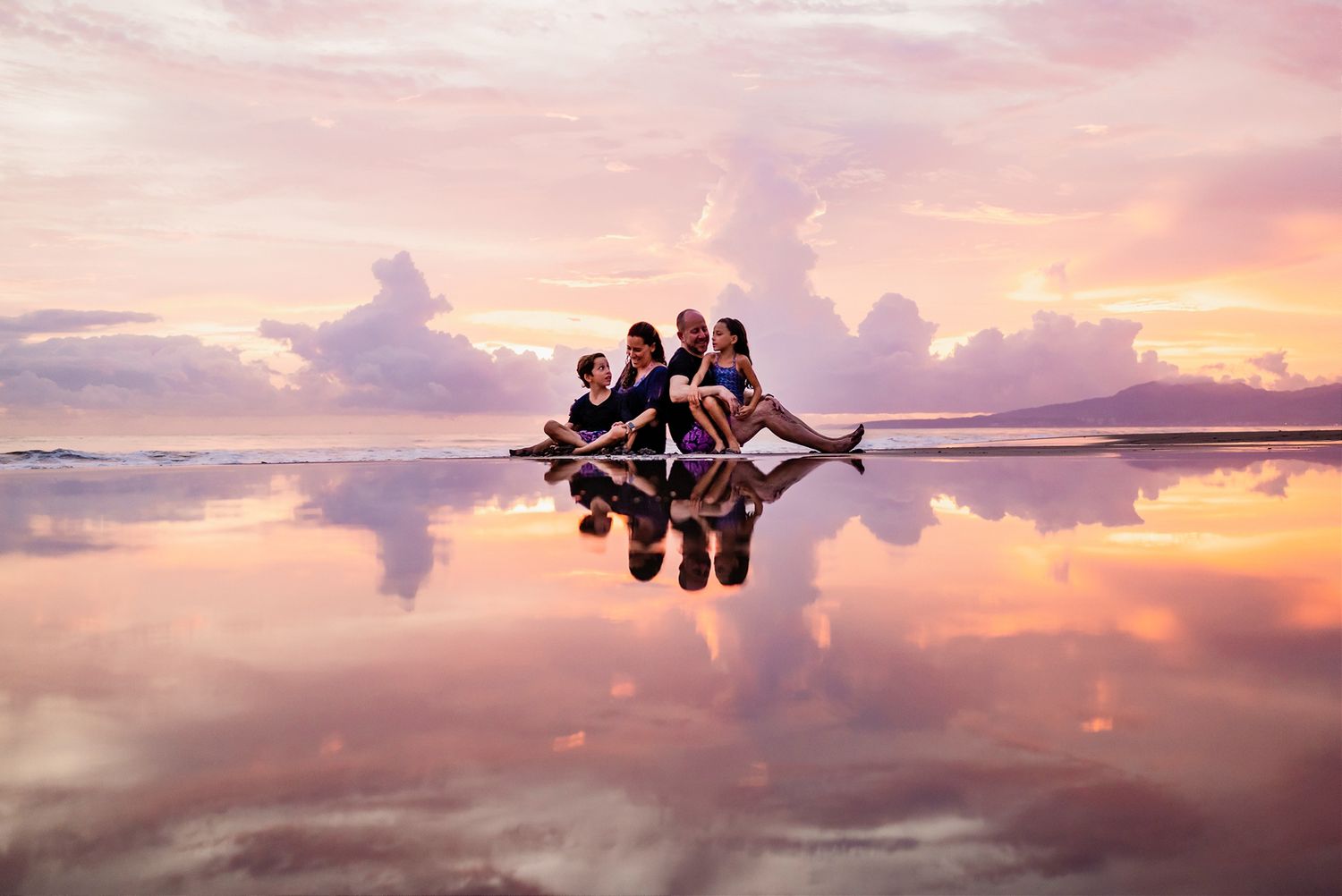 Silhouetted group sits on reflective salt flats during vibrant pink and orange sunset.