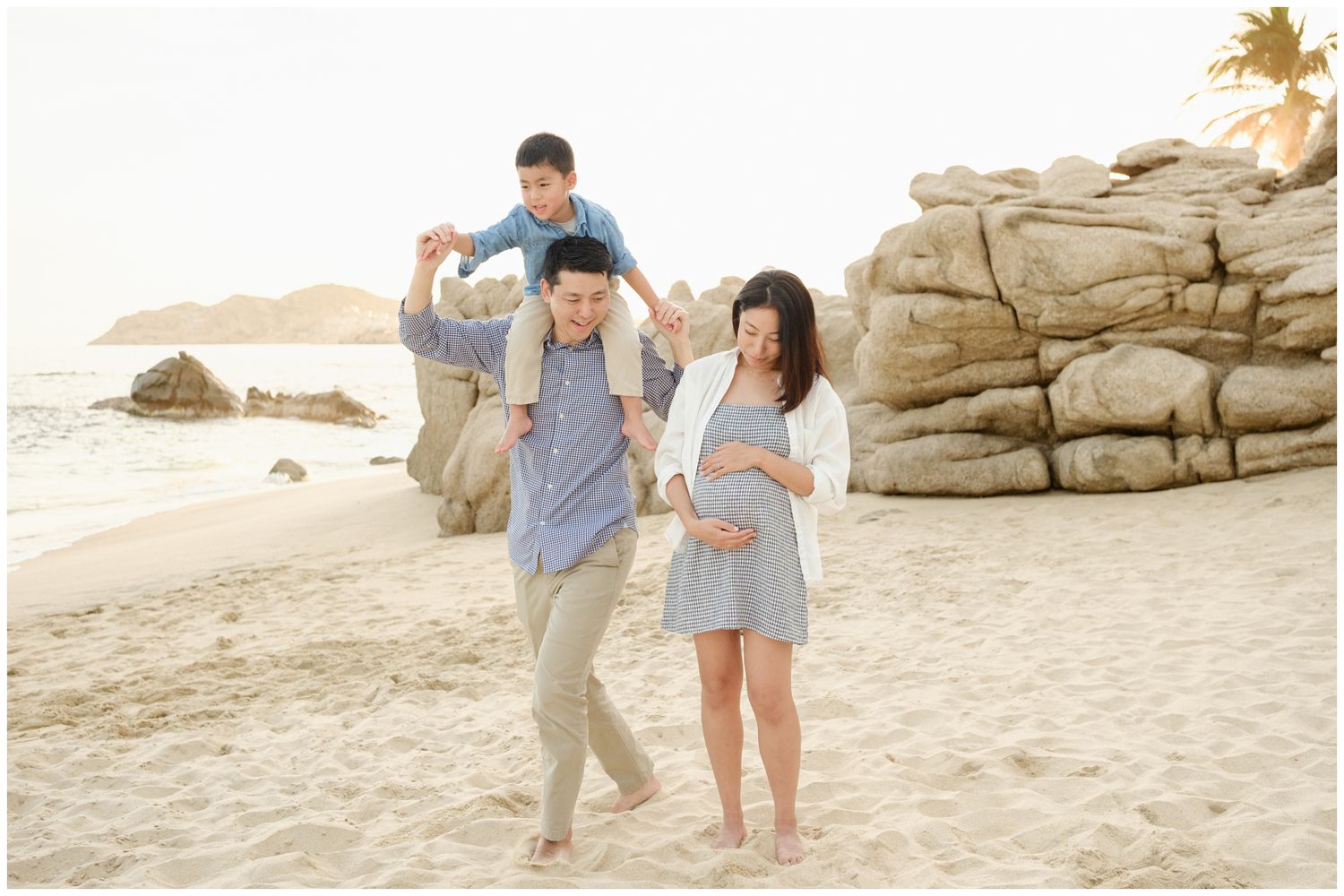 Two people in casual attire walk along a rocky beach path on a sunny day near the ocean.
