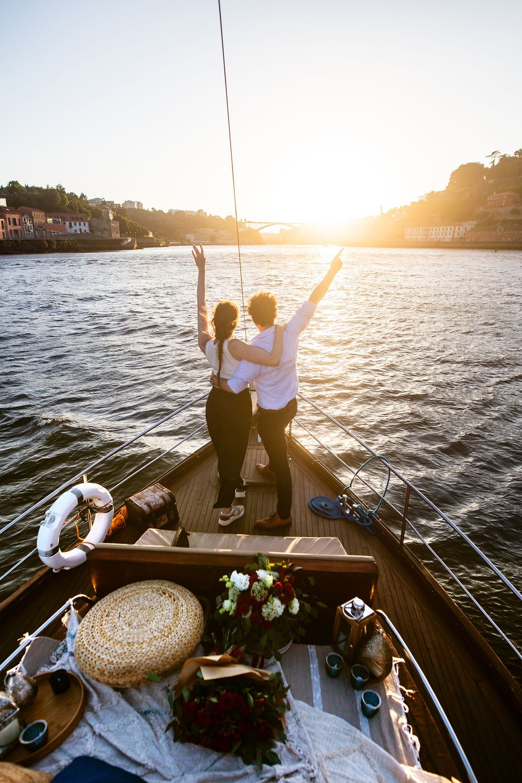 Couple standing on sailboat bow with arms raised watching golden sunset over river with picnic setup on deck.