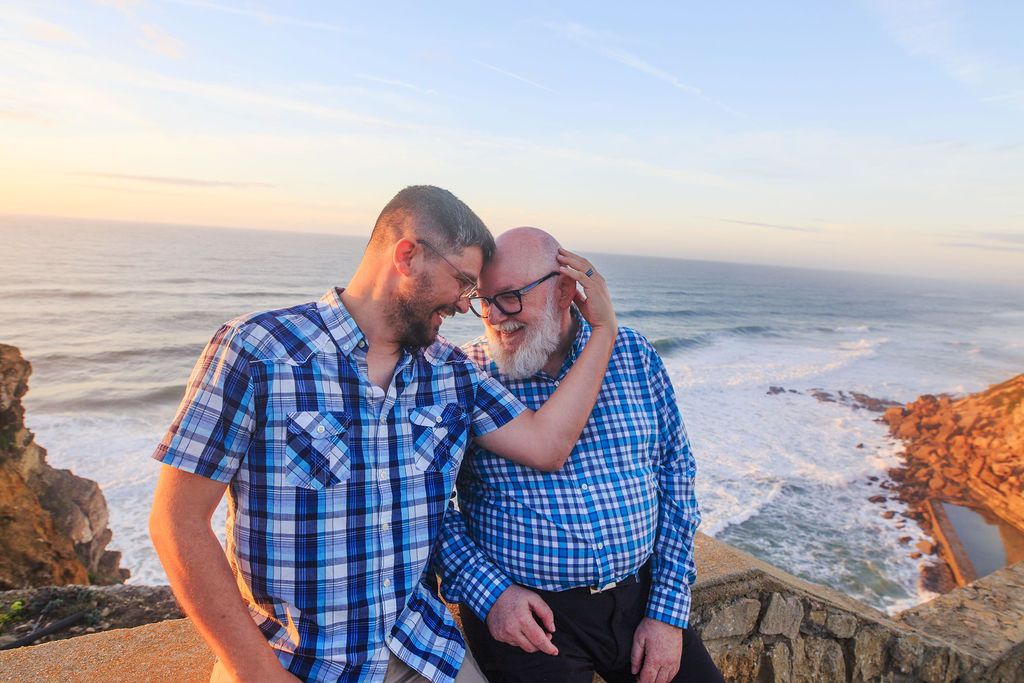 Two men in matching blue plaid shirts share a tender moment on a rocky coastal overlook at sunset.