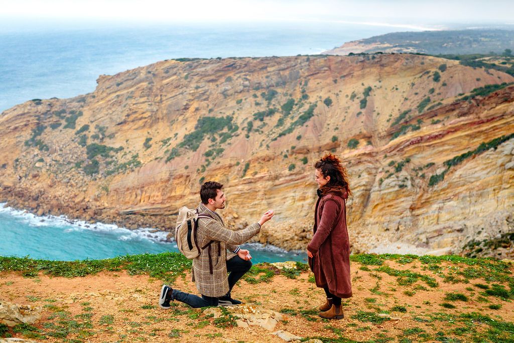 Man proposes to woman on scenic coastal cliffside with stunning ocean and rocky hillside views.