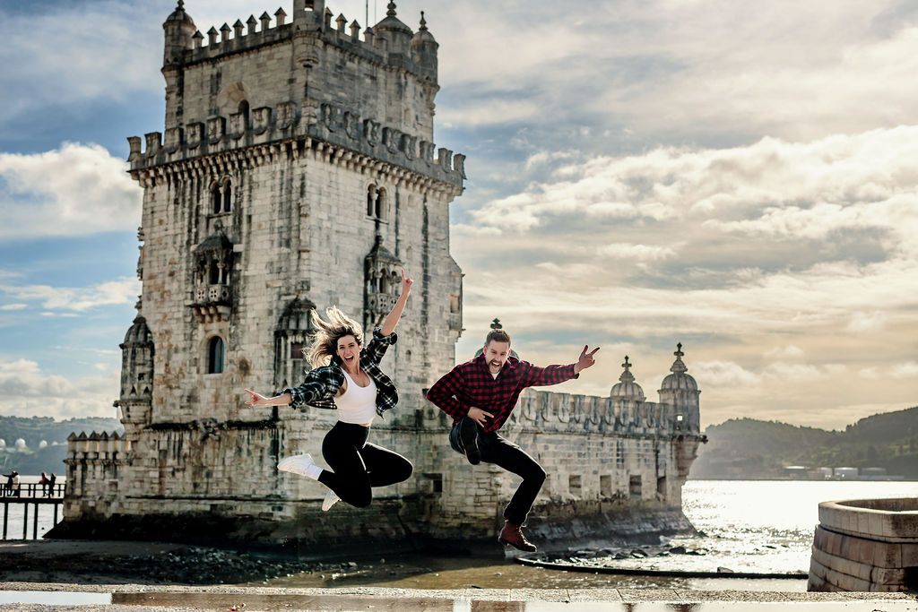 Two people jumping joyfully in front of the historic Belem Tower in Lisbon, Portugal on a cloudy day.
