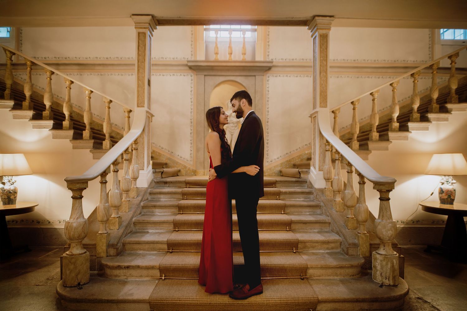 A couple shares an intimate moment on an elegant marble staircase in a historic building with warm sepia lighting.