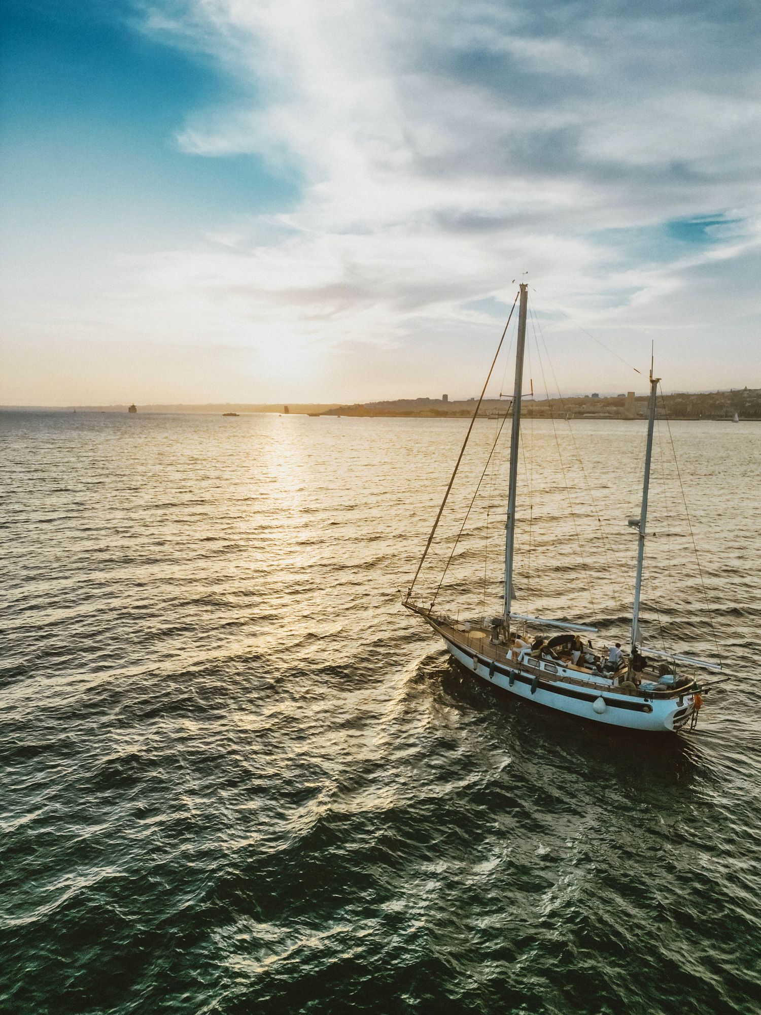 A sailboat navigates calm waters during golden sunset with dramatic clouds and shimmering reflections on the ocean.