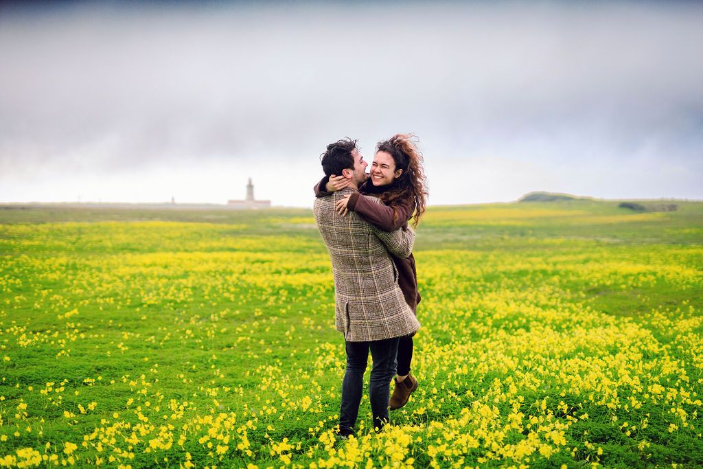 Couple embracing joyfully in a vibrant yellow flower field under a cloudy sky.