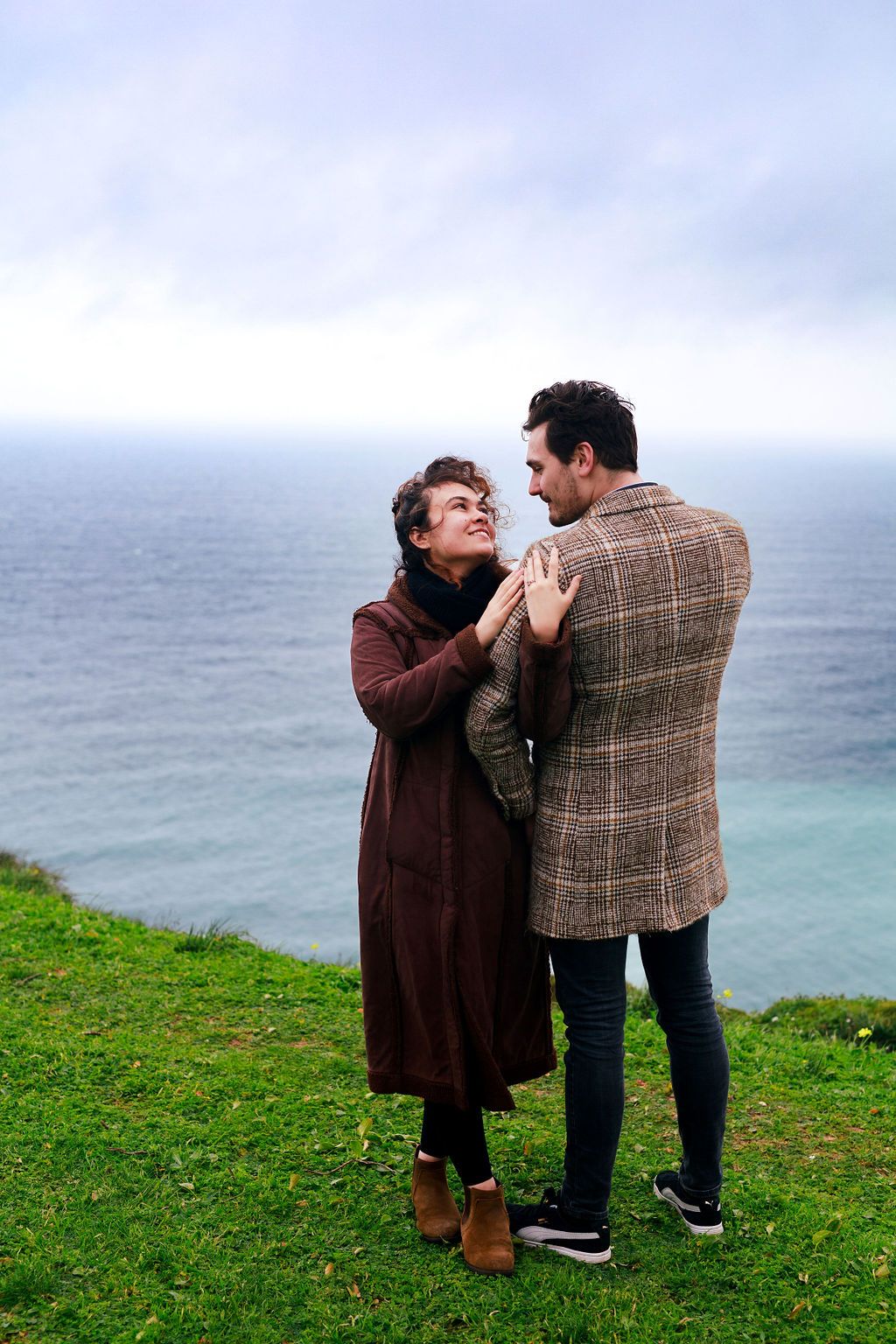 Couple holding hands on a coastal cliff, dressed in autumn coats, sharing a romantic moment with the ocean in the background.