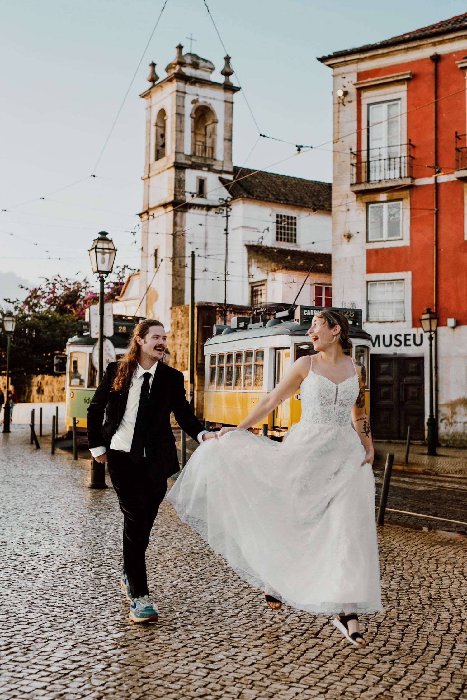 A couple in wedding attire dances on cobblestone streets of Portugal with a historic church tower and red building in background.