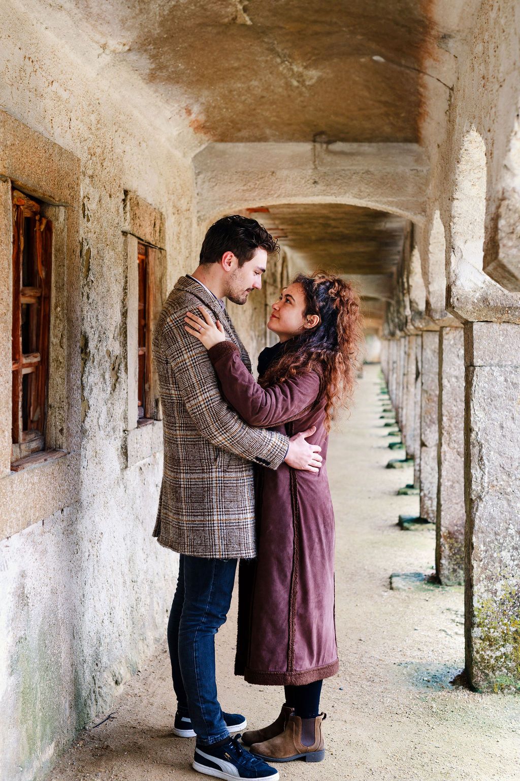 Romantic couple embracing in a rustic stone corridor, wearing stylish autumn coats, sharing an intimate moment.