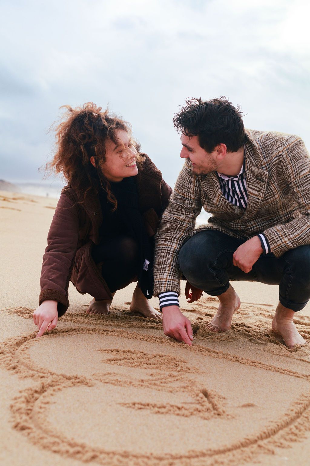 Couple crouching on a sandy beach, smiling at each other while drawing a heart shape in the sand.
