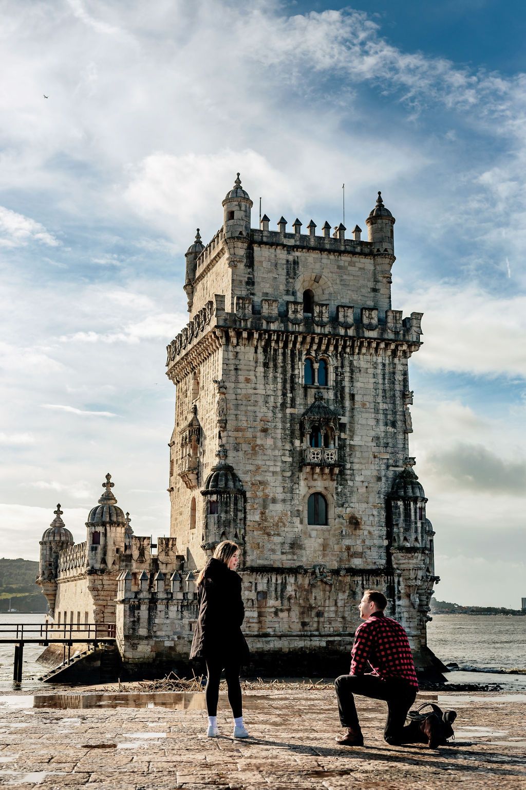 Man proposing to woman on one knee at Belem Tower, Lisbon, Portugal, with river and cloudy sky backdrop.