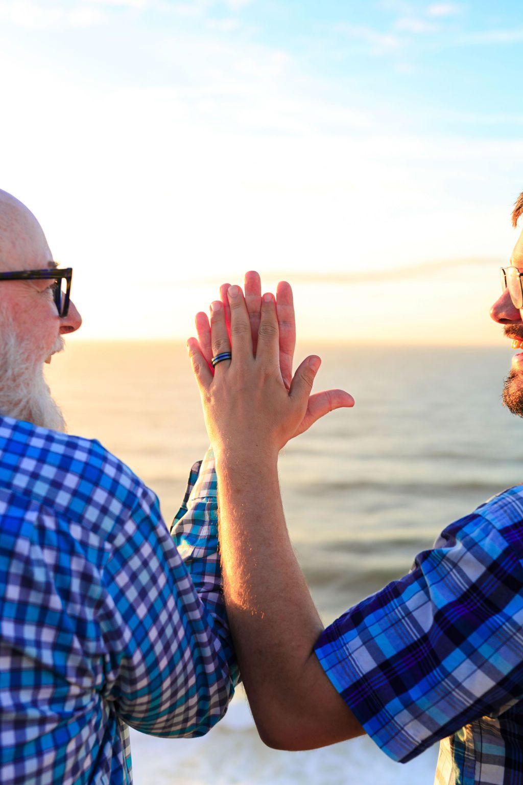 Two men holding hands up near ocean at sunset, wearing plaid shirts and glasses, with wedding rings visible.