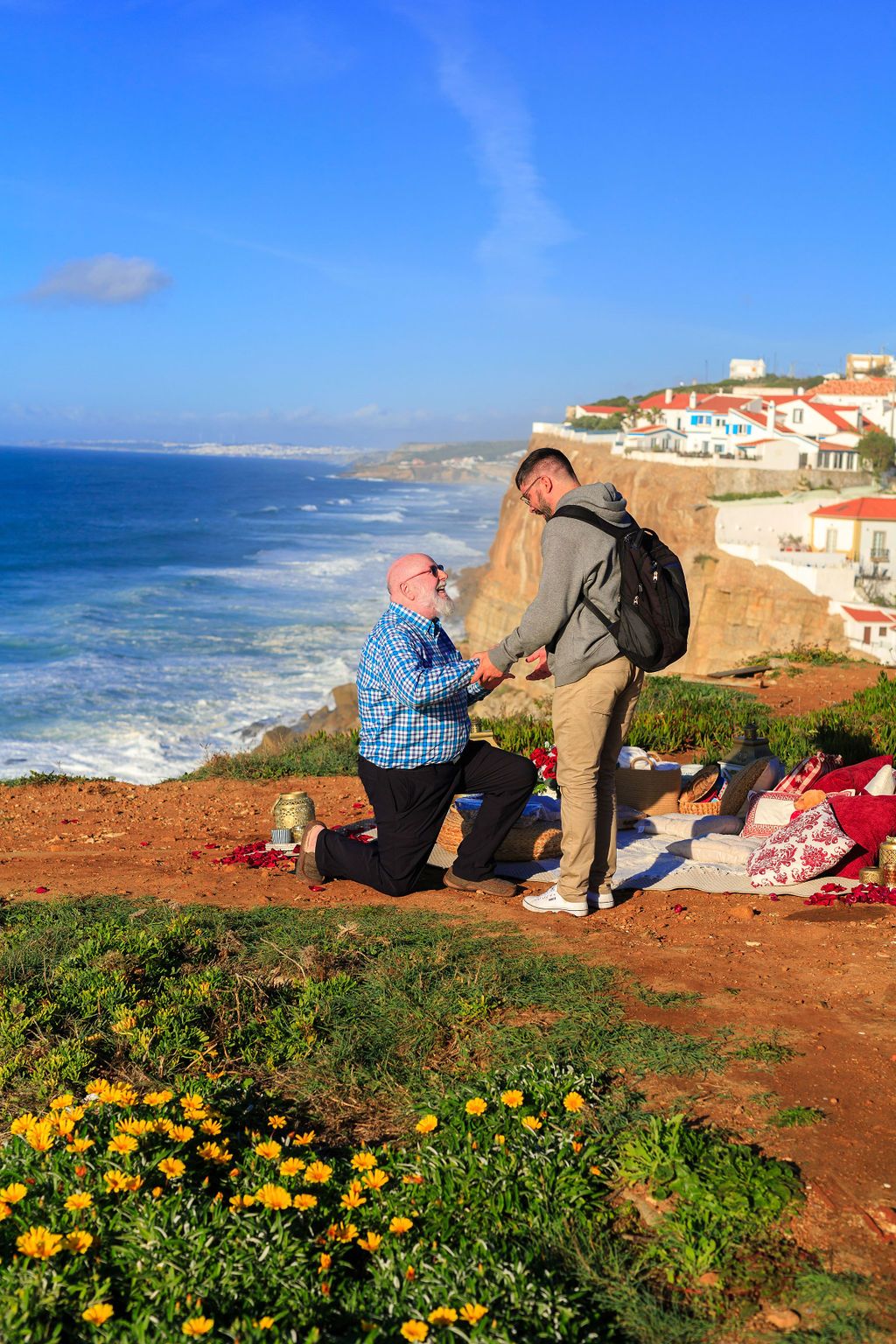 Older man proposes on one knee to partner on scenic coastal cliff overlooking ocean and village in Portugal.