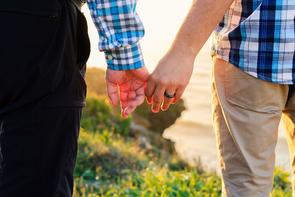 Couple holding hands outdoors near greenery and water, wearing casual clothing in warm sunlight.