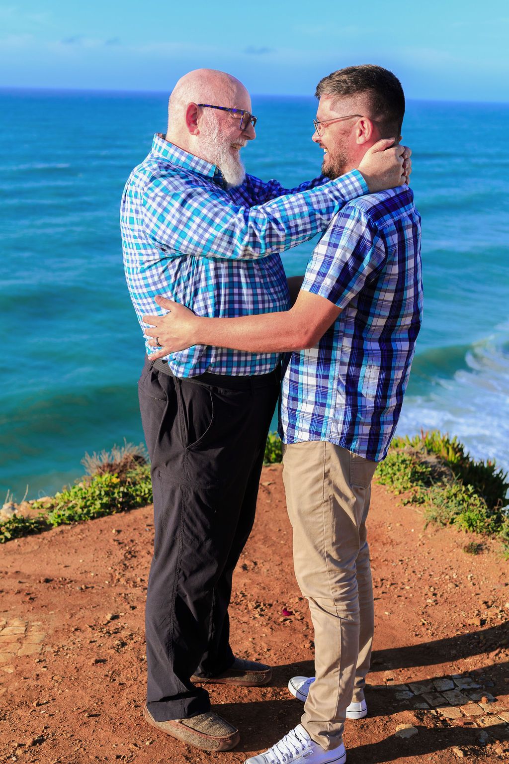 Two men in plaid shirts embrace on a red cliff overlooking a turquoise ocean on a sunny day.