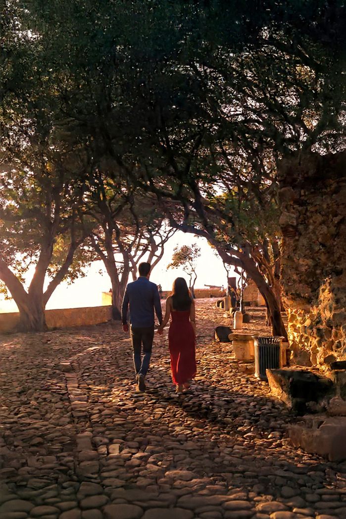 A couple walks hand in hand along a cobblestone path under trees at sunset creating a romantic silhouette.
