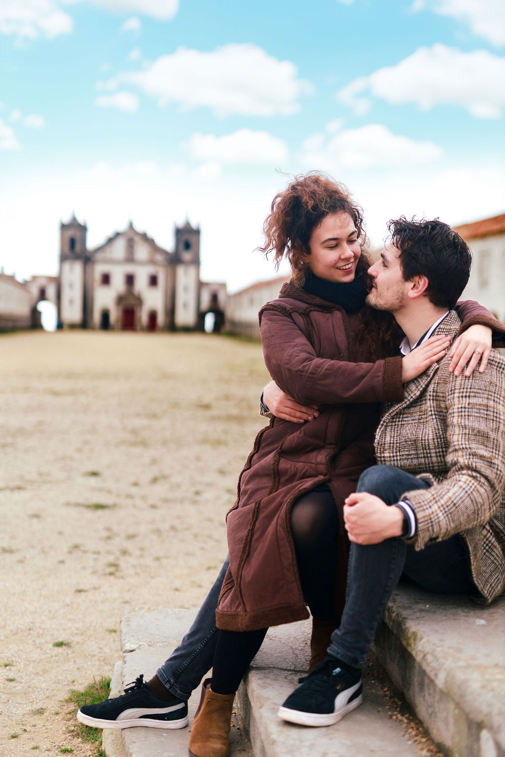 Romantic couple embracing on stone steps with a historic church in the background on a cloudy day.