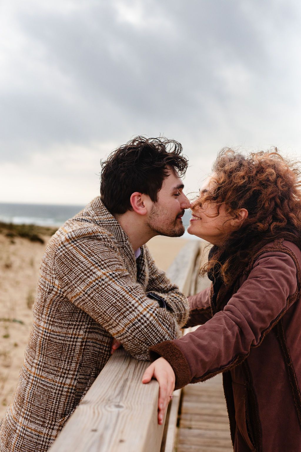 Romantic couple sharing a kiss on a wooden boardwalk by the beach on a cloudy day.
