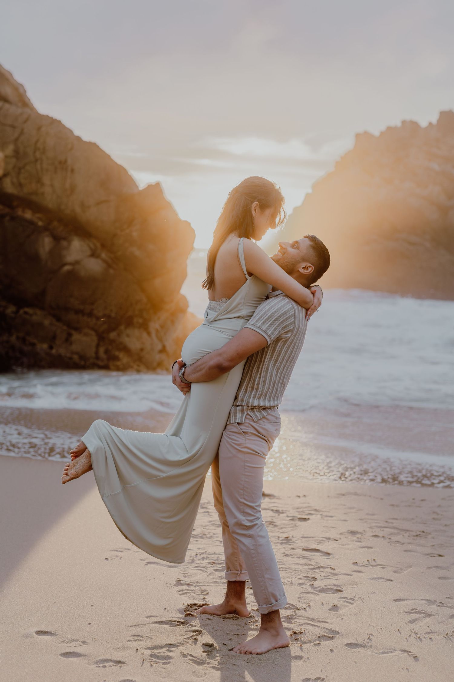Couple embracing on a sandy beach at golden sunset, with rocky cliffs and ocean waves in the background.