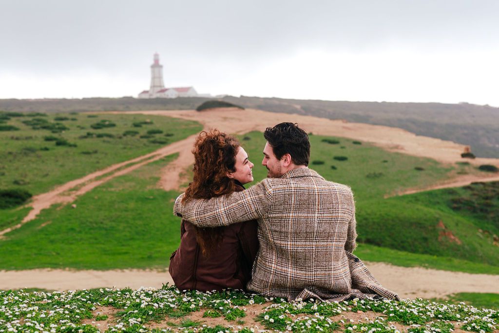 Couple sitting together on a grassy hillside overlooking a lighthouse on a cloudy day.
