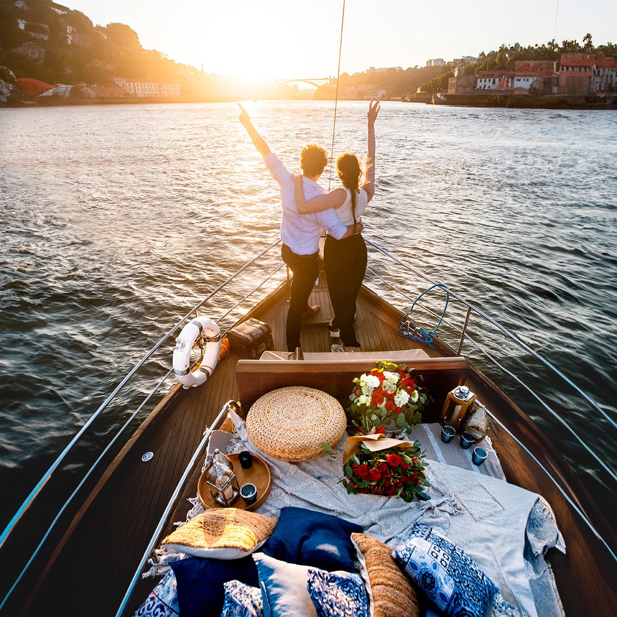 A romantic sunset boat ride on calm waters with picnic essentials including wine, flowers, and a straw hat laid out.