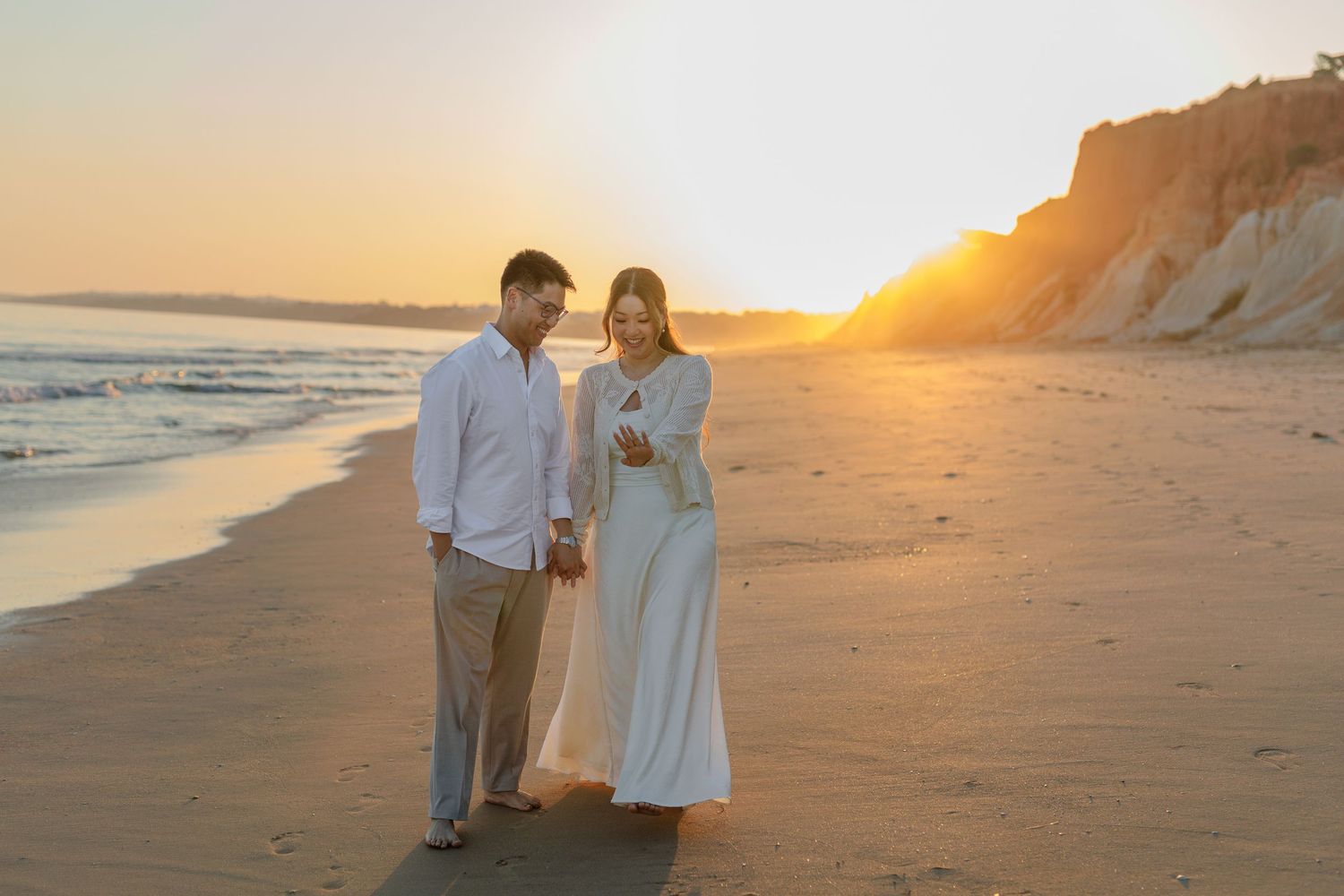 Couple in white attire walking on a sandy beach at golden sunset with cliffs in the background.