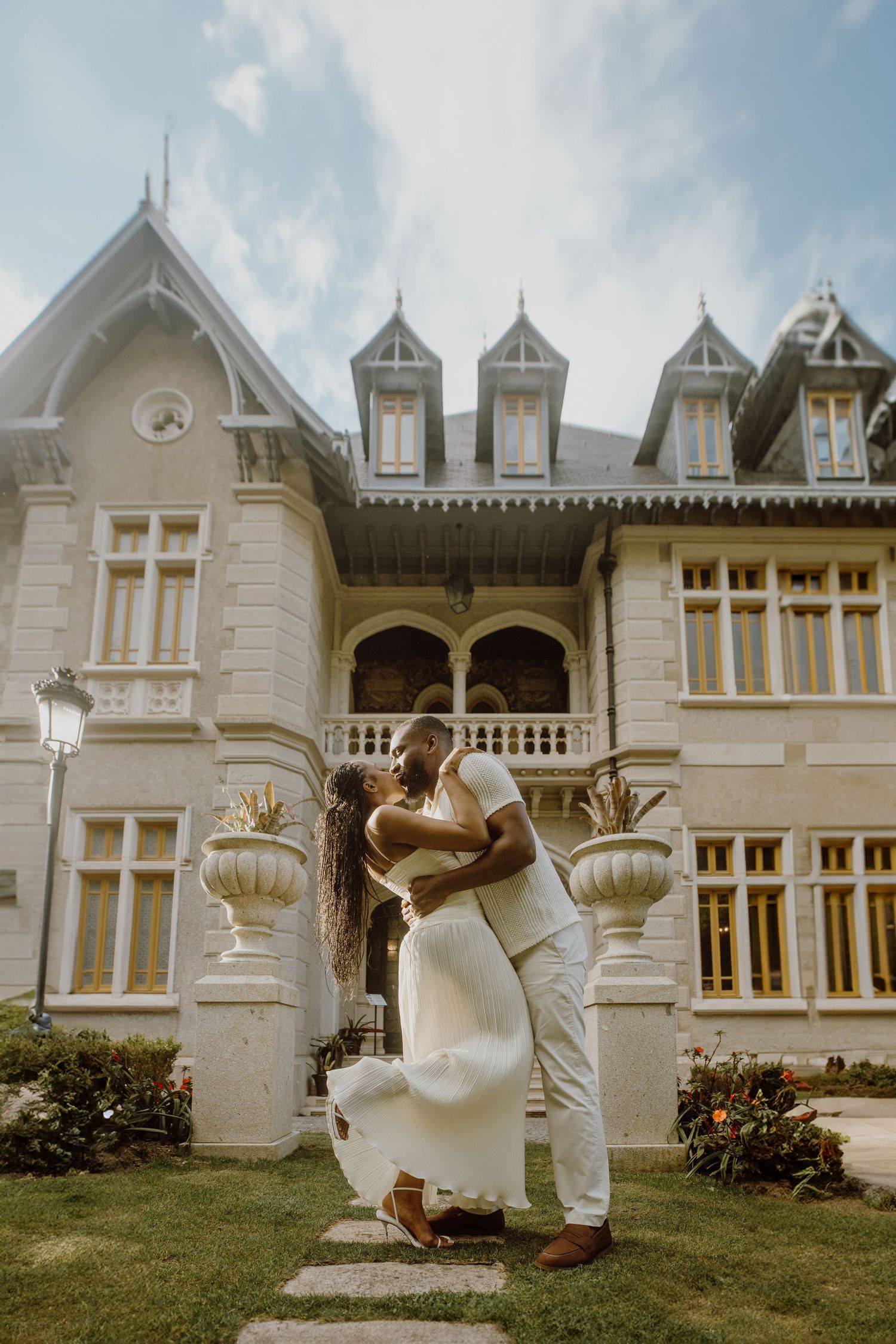 Couple sharing a romantic kiss in front of an elegant white mansion on a sunny day.