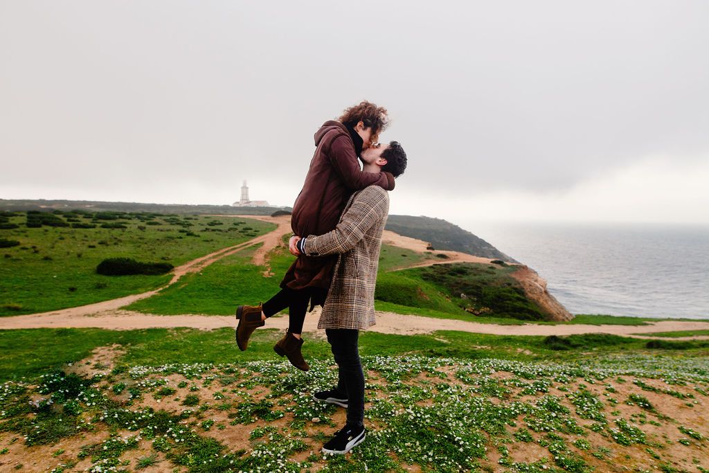 Couple embracing romantically on a scenic coastal cliff with green hills and ocean views on a misty day.