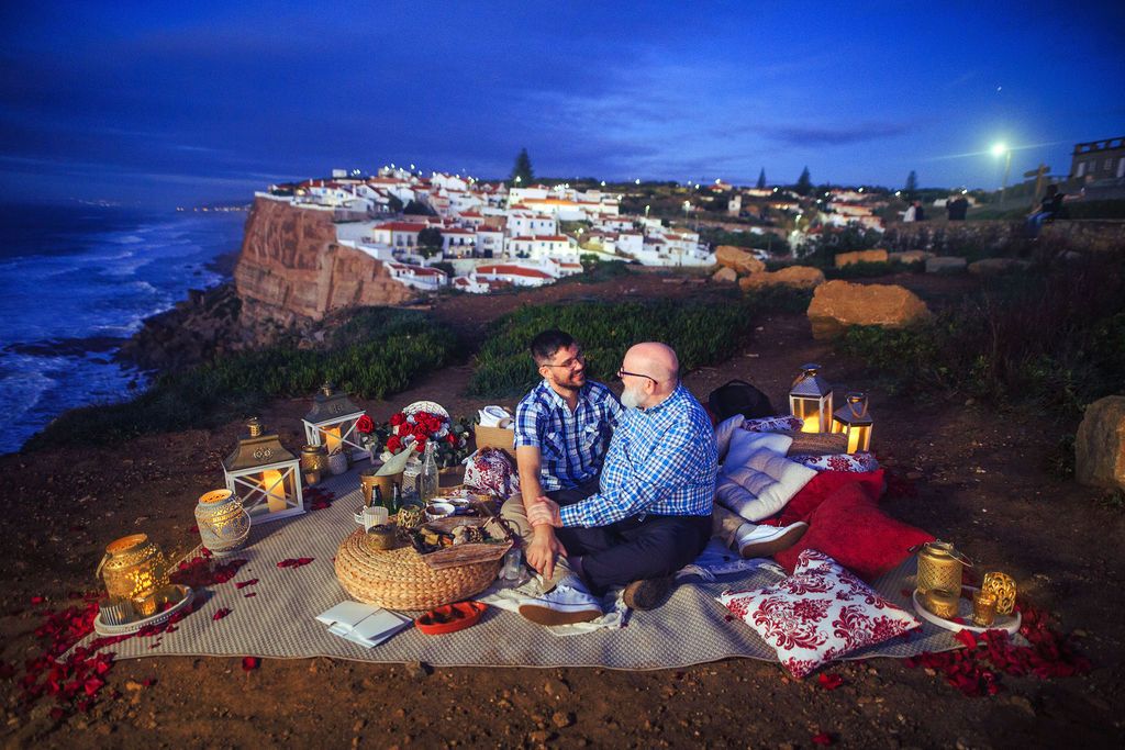 Couple enjoying a romantic cliffside picnic at dusk with ocean views and a coastal village backdrop.