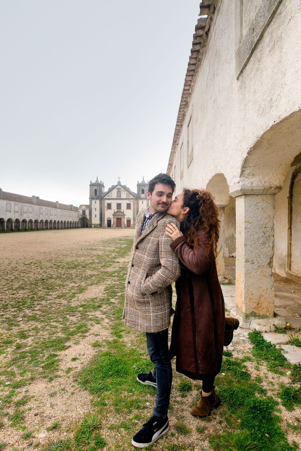 Couple sharing a romantic kiss outside a historic European church courtyard, dressed in stylish winter coats.