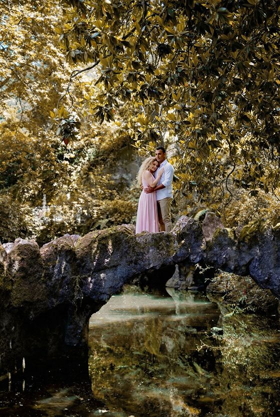 A couple embraces on a mossy stone bridge under golden-lit tree branches reflecting in still water below.