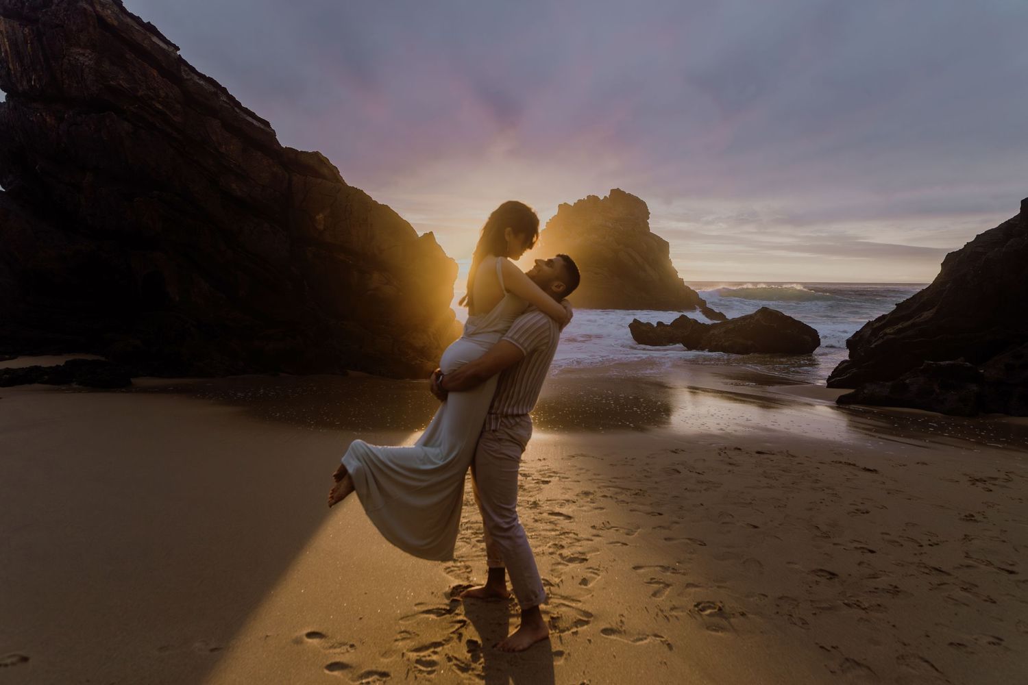 Couple embracing on a sandy beach at sunset, surrounded by dramatic rock formations and golden light.