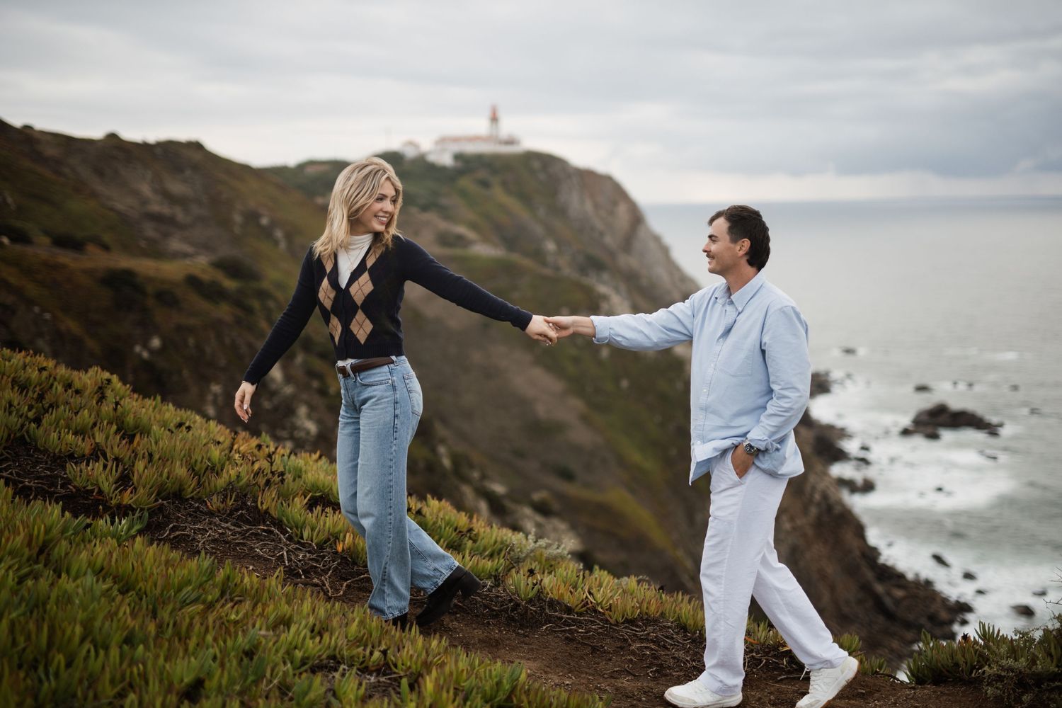 Couple holding hands on a scenic coastal cliff with a lighthouse and ocean waves in the background.