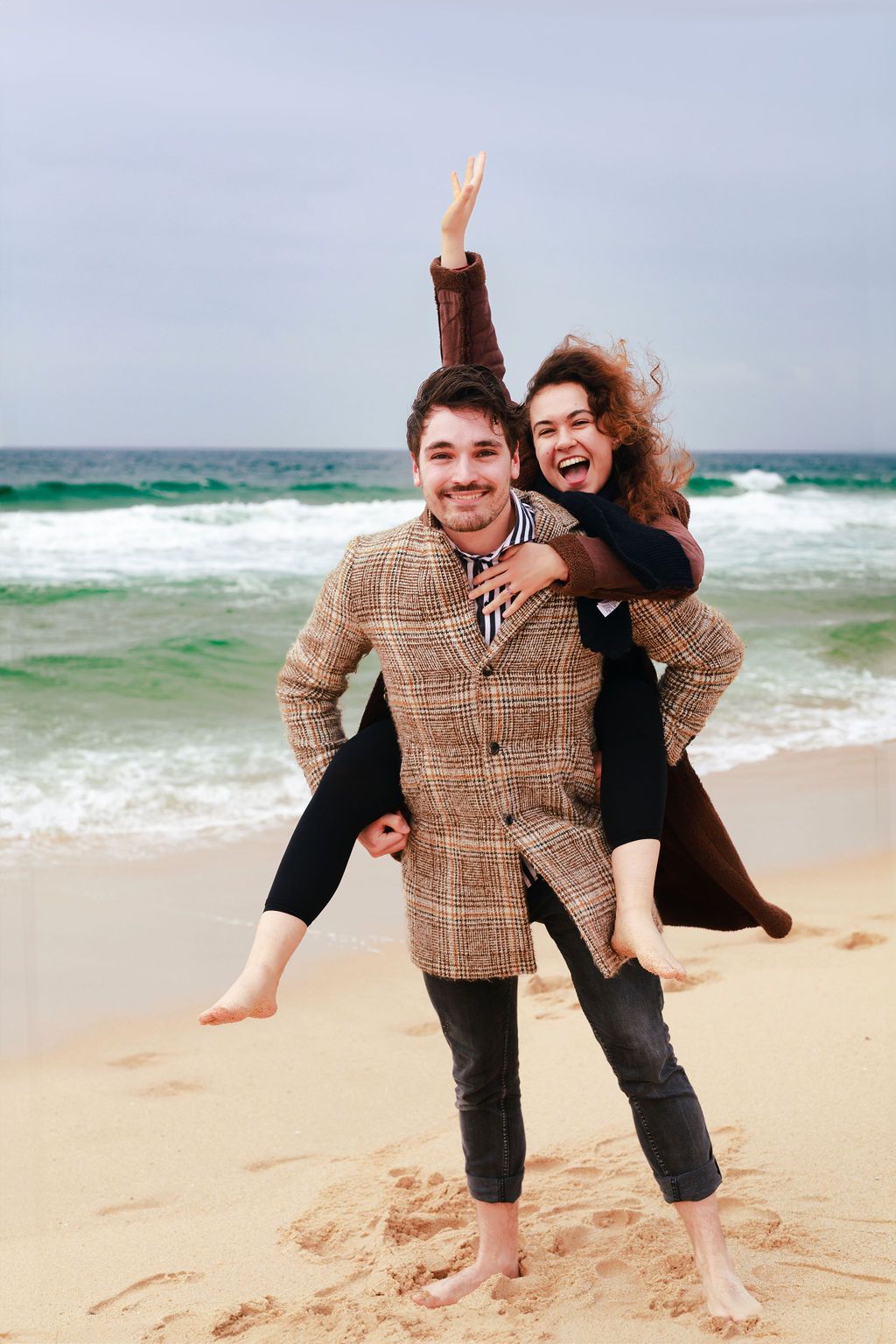 Happy couple enjoying a piggyback ride on a sandy beach, with ocean waves in the background on a cloudy day.
