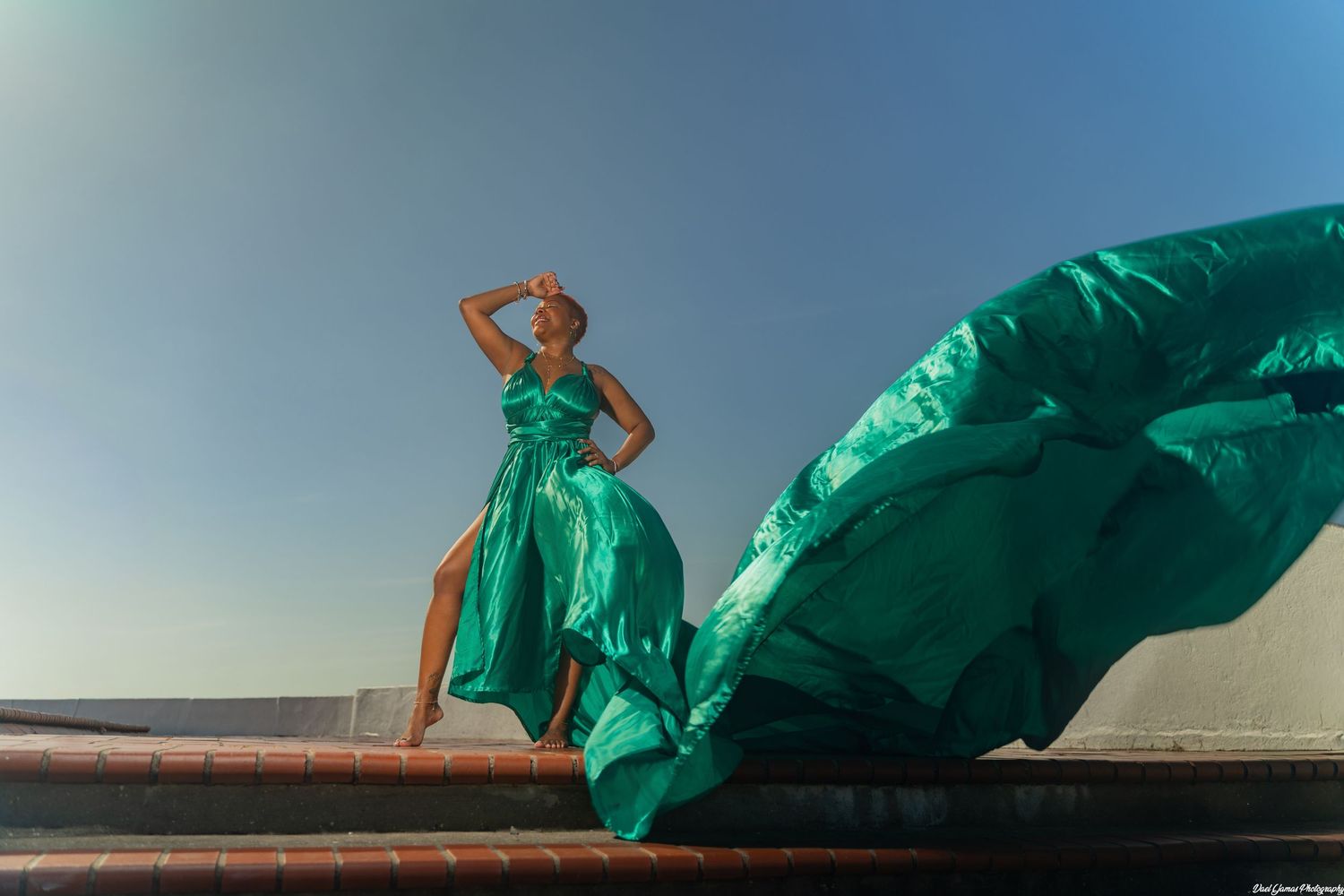 A flowing emerald green dress billows dramatically against a clear sky as someone poses on a rooftop.