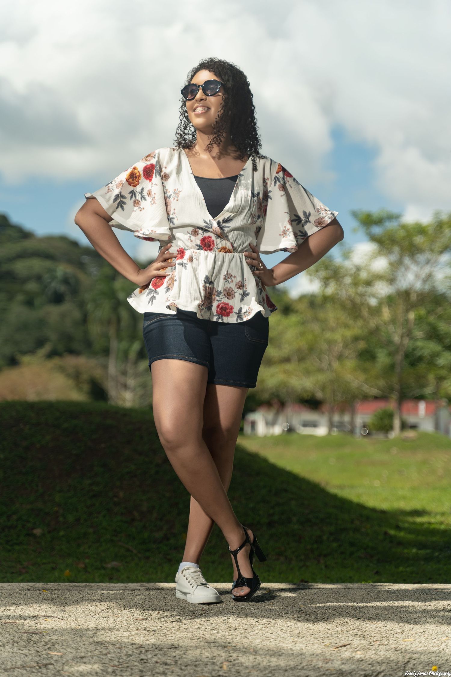 A person in a floral wrap top and black shorts poses confidently outdoors on a sunny day with greenery in background.