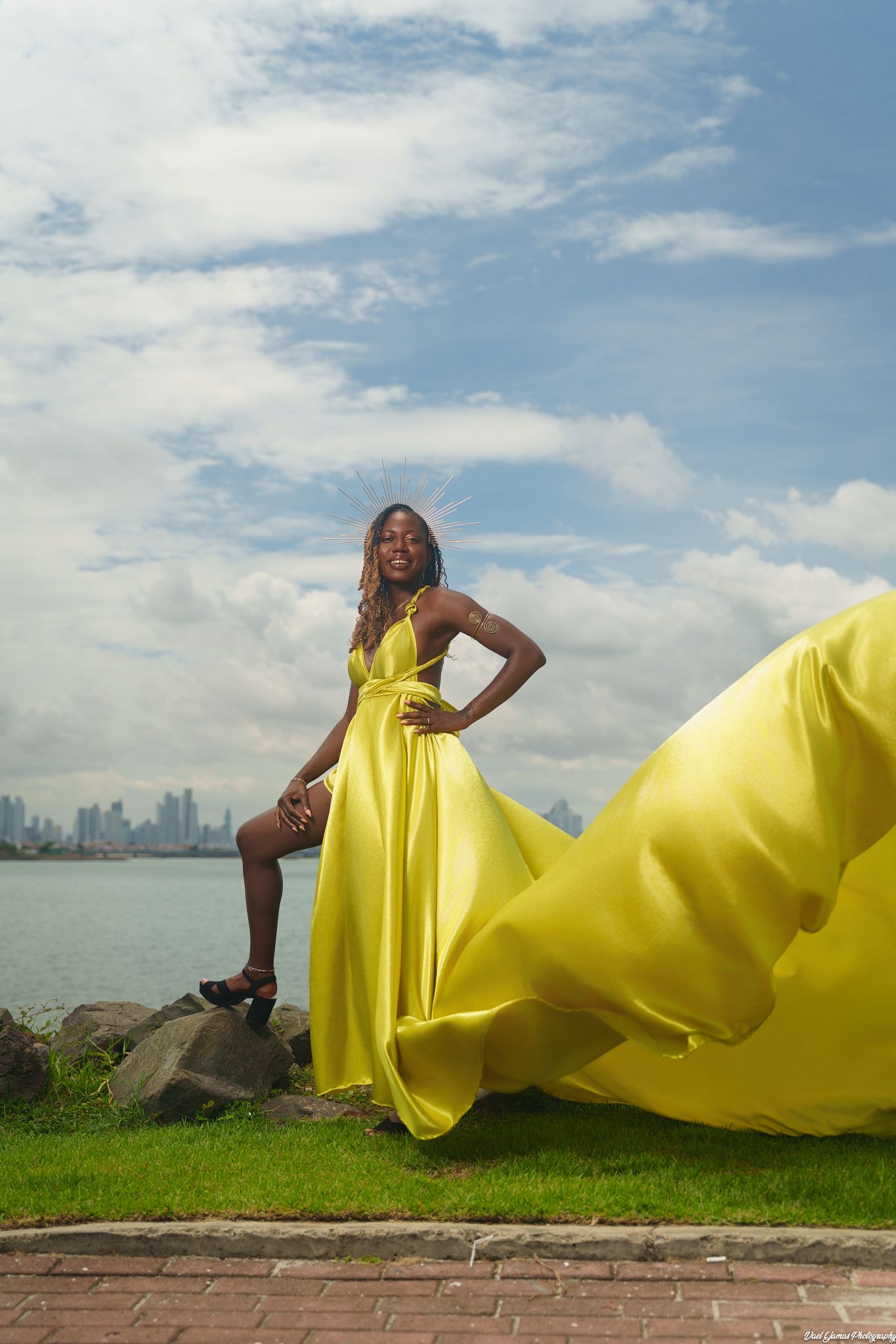 A flowing yellow gown billows dramatically against a waterfront cityscape backdrop on a sunny day.