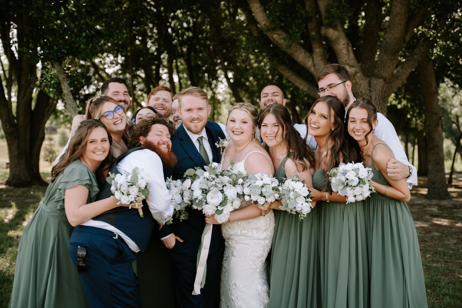 Wedding party in sage green bridesmaid dresses pose together with white floral bouquets under trees.