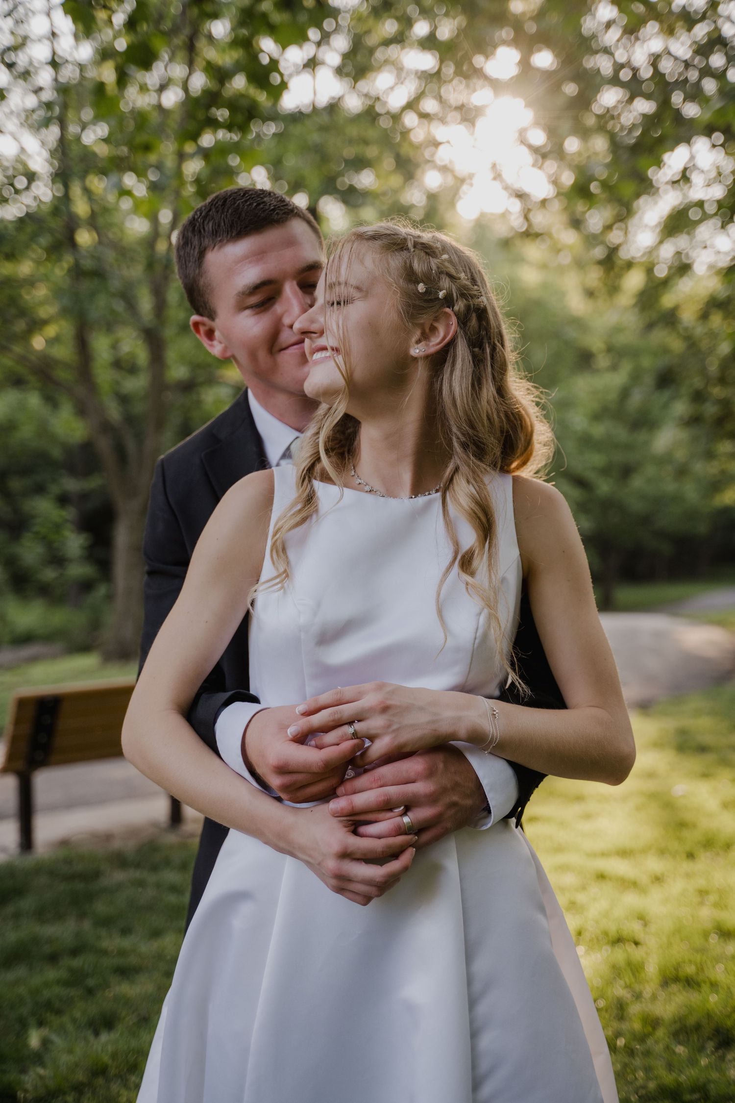 A romantic couple embraces in a sun-dappled garden setting wearing formal attire.
