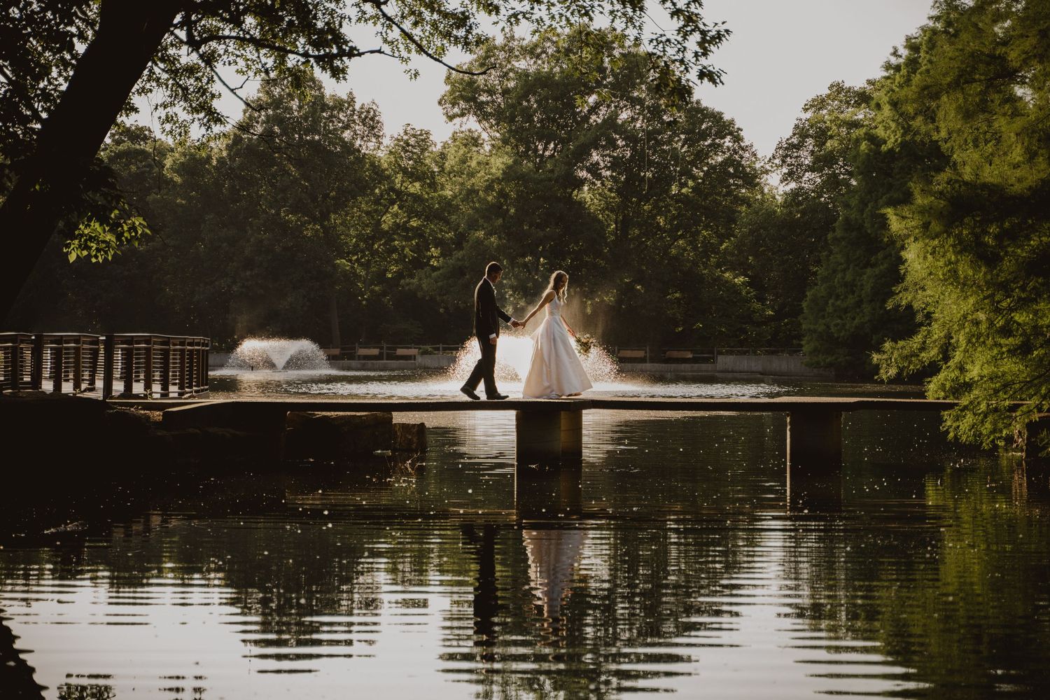A romantic wedding couple poses in a boat on a serene lake surrounded by lush green trees at sunset.